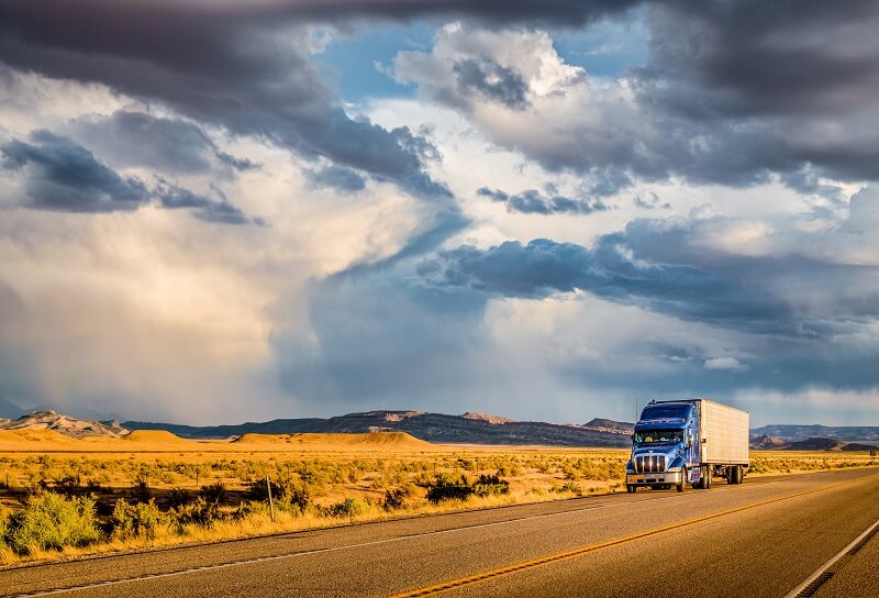 A tractor trailer driving on a desert road with dunes and mountains in the background.